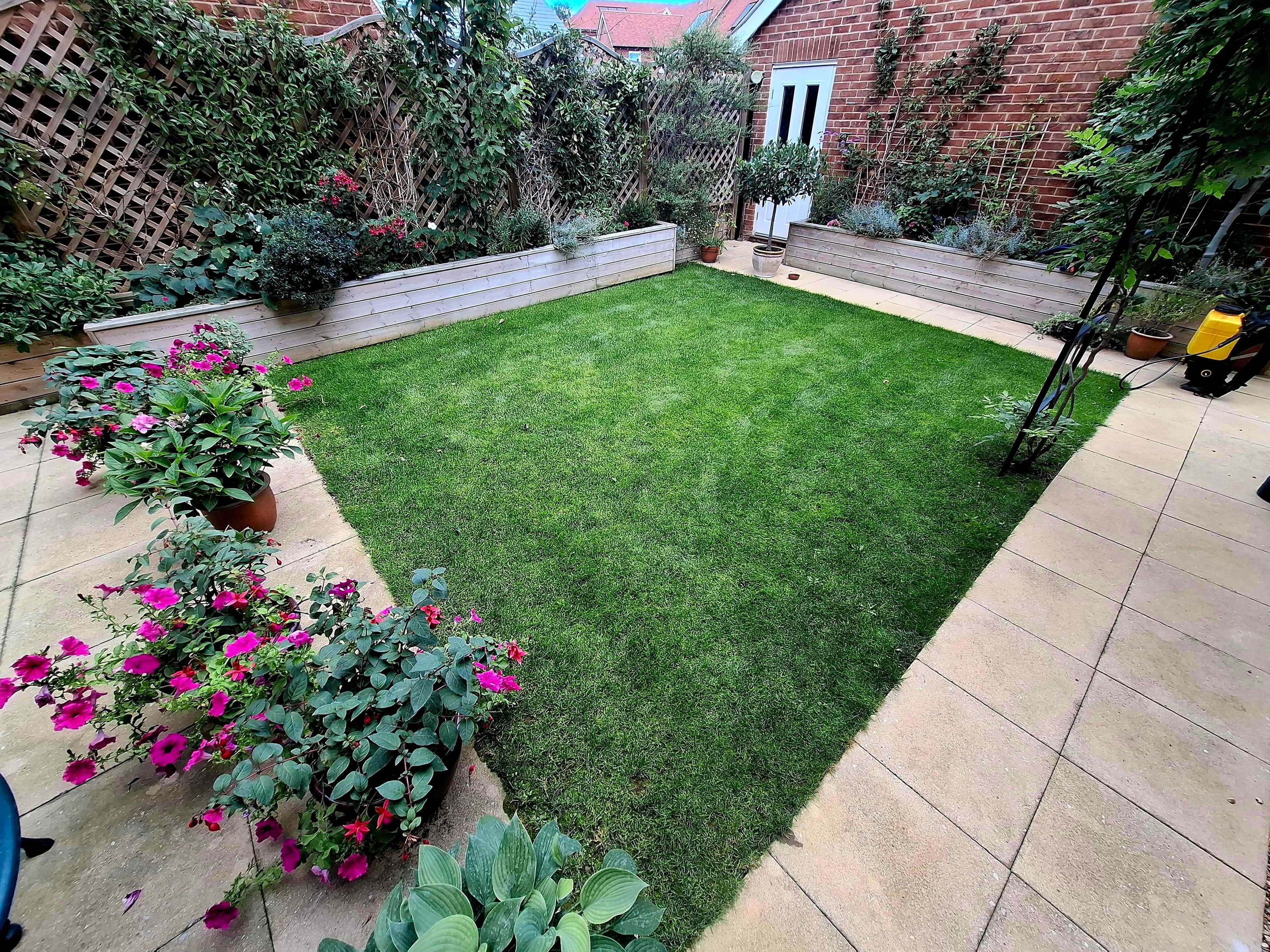 Picture of a garden with a green, lush lawn with flowers and a patio around its perimeter.